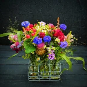 Cute basket garden formed from mini milk bottles, containing an assortment of pretty flowers which may include beauties like roses, ranunculus, veronica and wax flower.